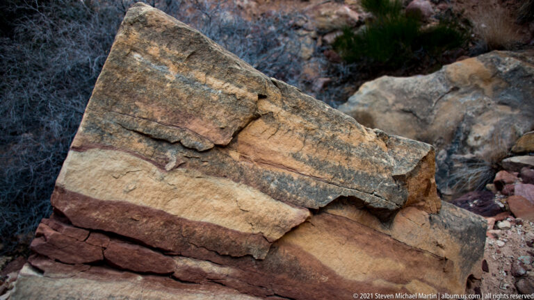 Angular, layered sandstone blocks sit among brush, with fractured edges and a dark weathered patch on the top slab.