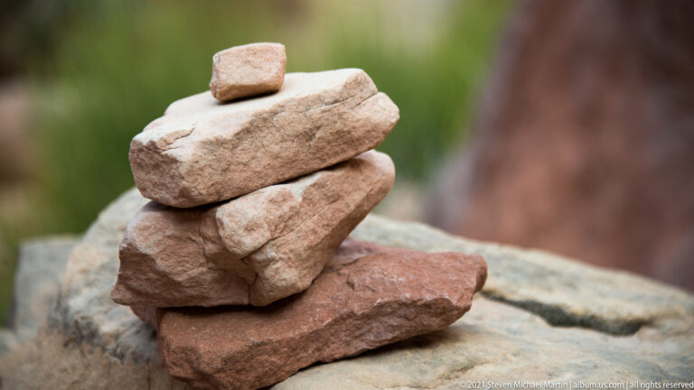 Flat tan and reddish stones form a small balanced cairn on a larger rock, with one rounded stone on top.