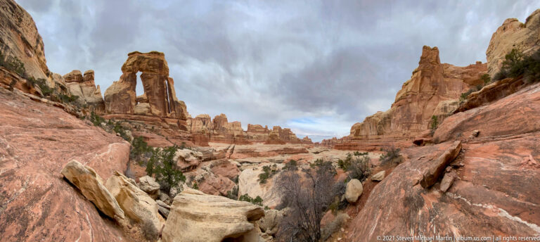 A distant Druid Arch stands above pale boulders and shrubs between layered canyon walls.