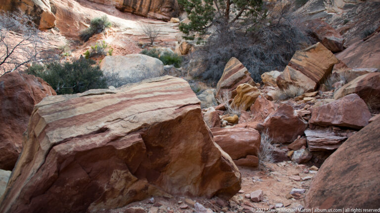A striped boulder and broken sandstone slabs fill a brushy canyon floor, with a narrow rocky route beyond.