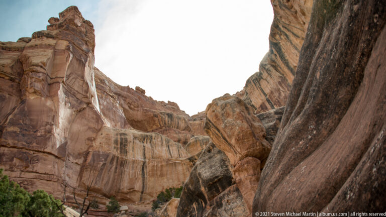 Steep sandstone walls enclose a narrow canyon, with boulders in the route and a pale opening of sky above.