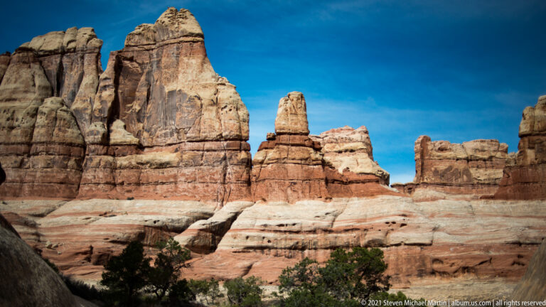 Sunlit sandstone towers and fins rise above a pale ledge, with a narrow pinnacle against a clear blue sky.