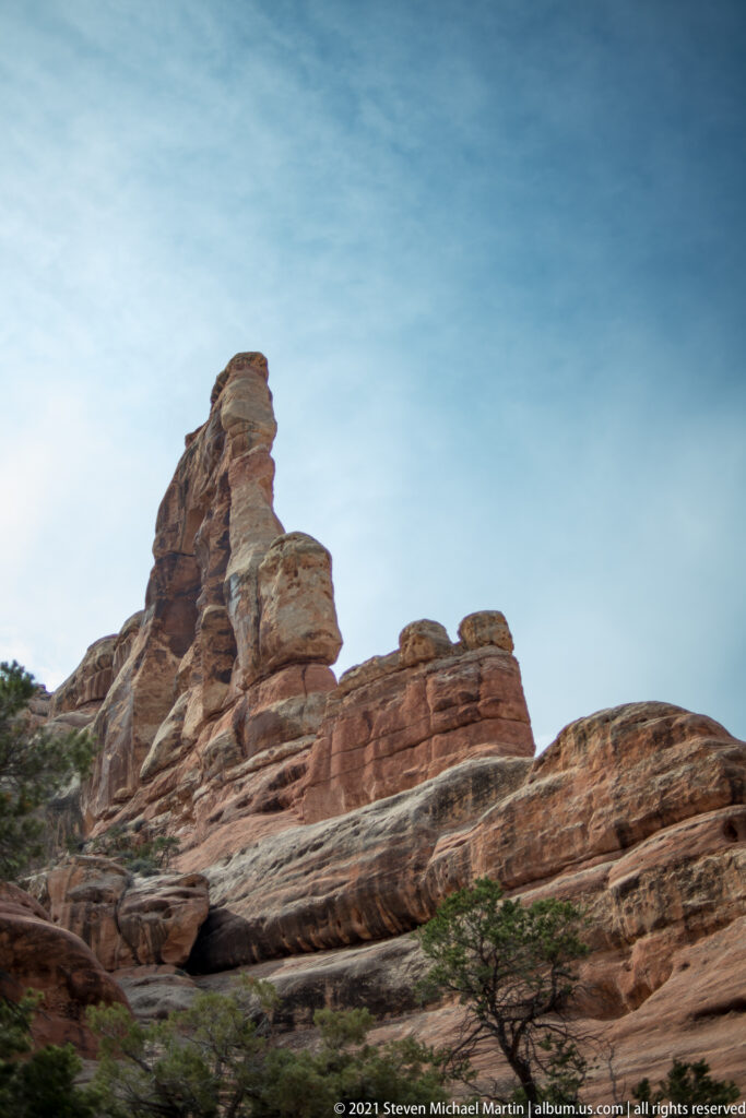 A low sandstone ridge rises to a leaning rock tower, with sparse shrubs along the slope beneath a pale sky.