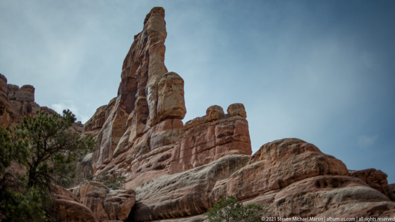 A tall narrow sandstone spire rises among rounded rock formations and sparse vegetation beneath a cloudy sky.