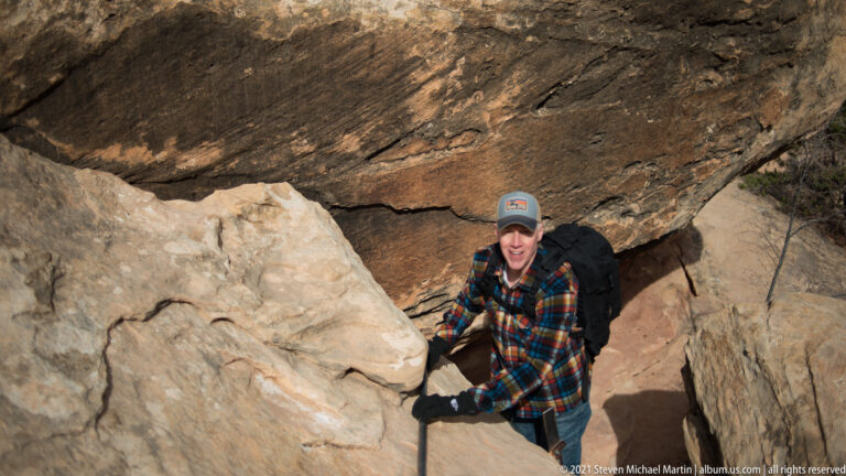 A hiker braces against sandstone while squeezing through a narrow gap beneath a dark overhang.