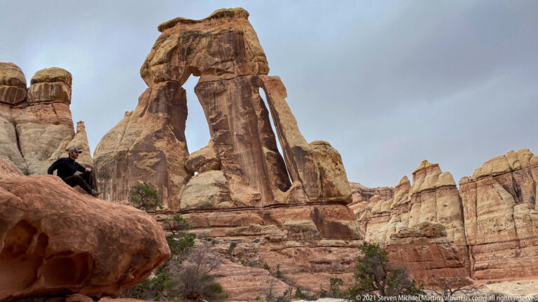 A seated hiker rests on a foreground boulder below Druid Arch, surrounded by tall sandstone towers.
