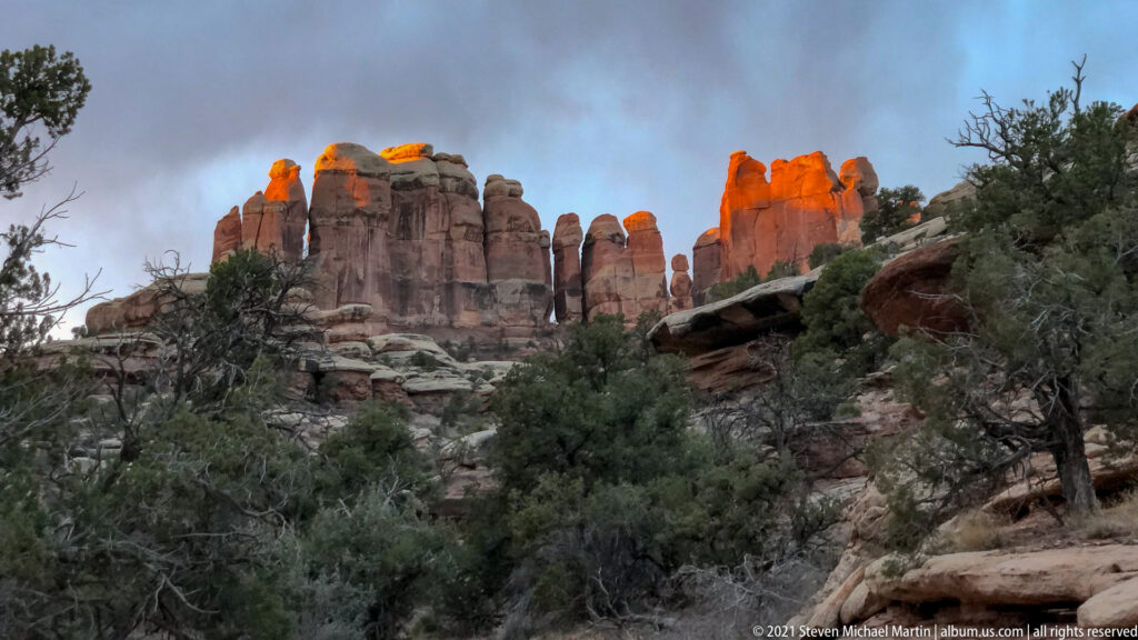 Orange sunlight illuminates the tops of red sandstone towers above shadowed shrubs and juniper.