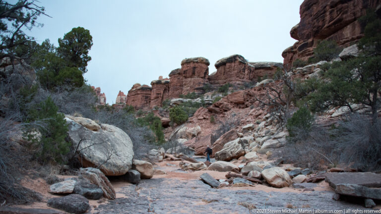 A small hiker walks away along a pale rocky wash between juniper and red sandstone towers.