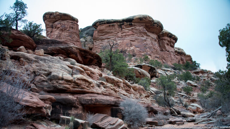 Rounded sandstone towers stand above sloping ledges and brush, with a dark wall at right.