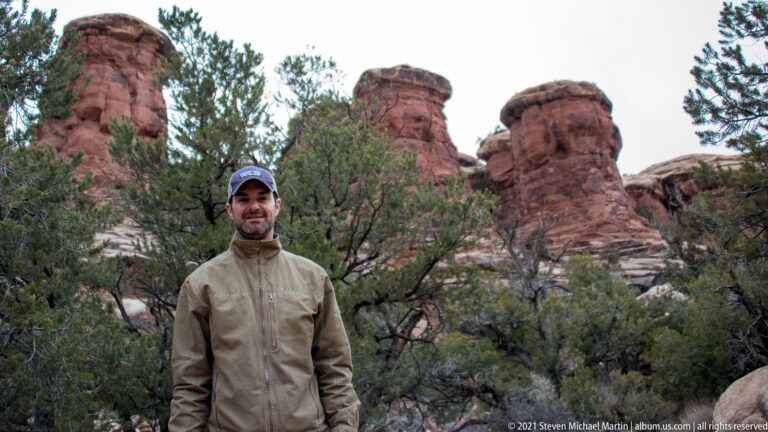 A man in a tan jacket and blue cap stands among junipers with red sandstone towers behind.
