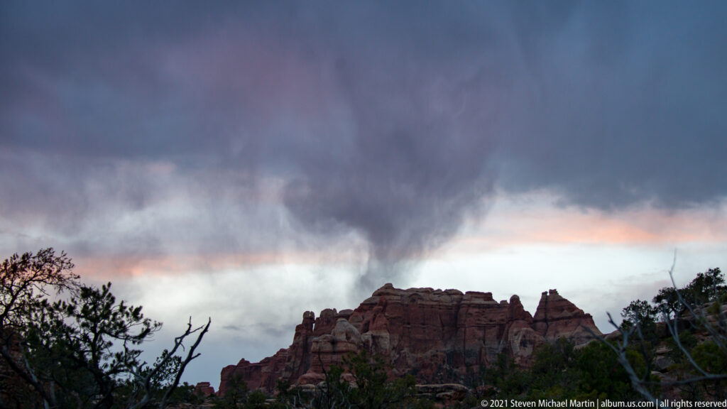 A dark storm cloud hangs above a distant sandstone skyline with a narrow warm band at the horizon.