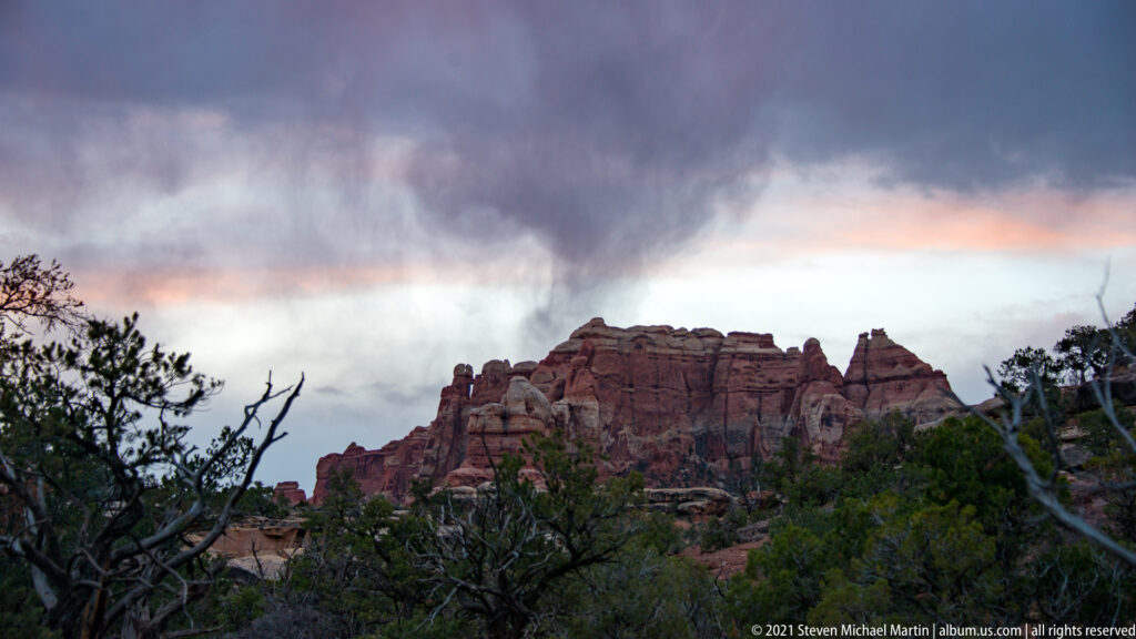A peach band of sunset light appears beneath a dark cloud above a jagged sandstone ridge.