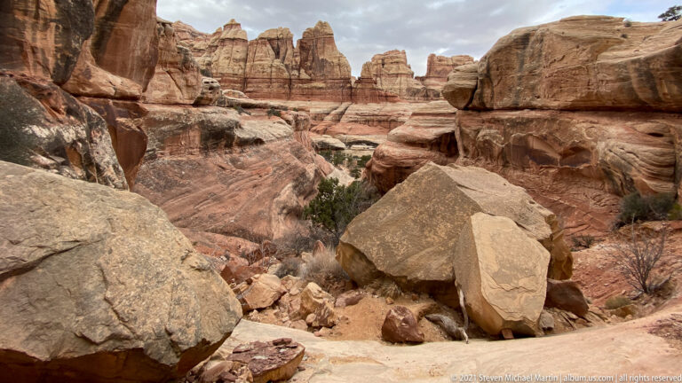 Pale boulders fill a dry wash below layered sandstone towers and a high wall at right.