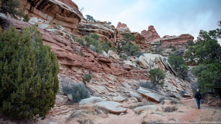 A hiker walks uphill on a pale rocky path beside layered sandstone and shrubs.