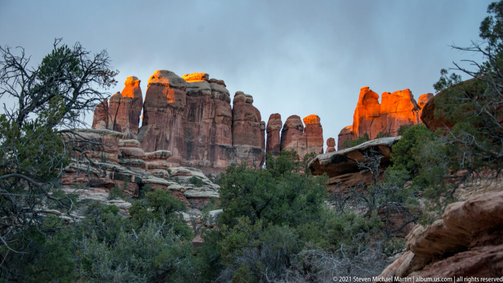 Orange sunlight strikes the tops of sandstone towers above a shadowed slope of juniper and brush.