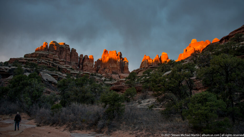 A small hiker walks toward orange-lit sandstone towers beneath a broad dark storm cloud.