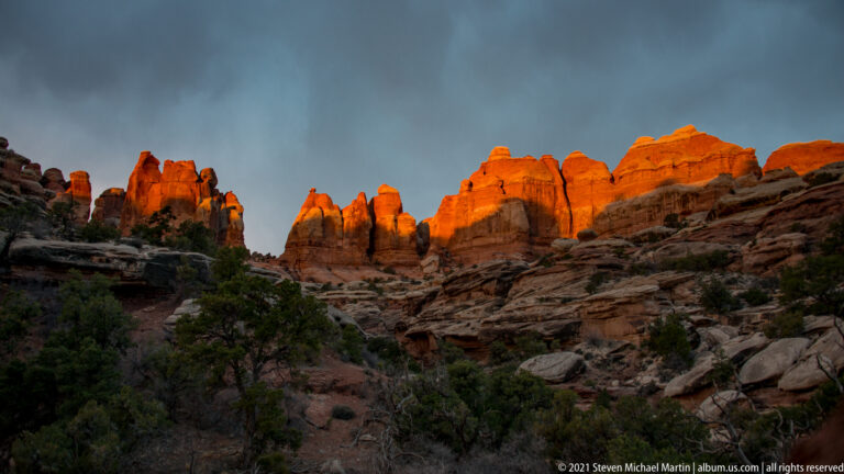 Orange-lit sandstone towers rise above shadowed sloping ledges beneath a narrow gray-blue sky.