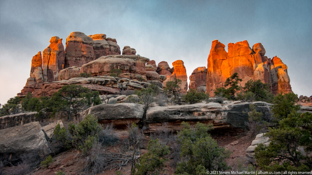 A continuous sandstone skyline catches orange sunset light above dark ledges and shrubs.