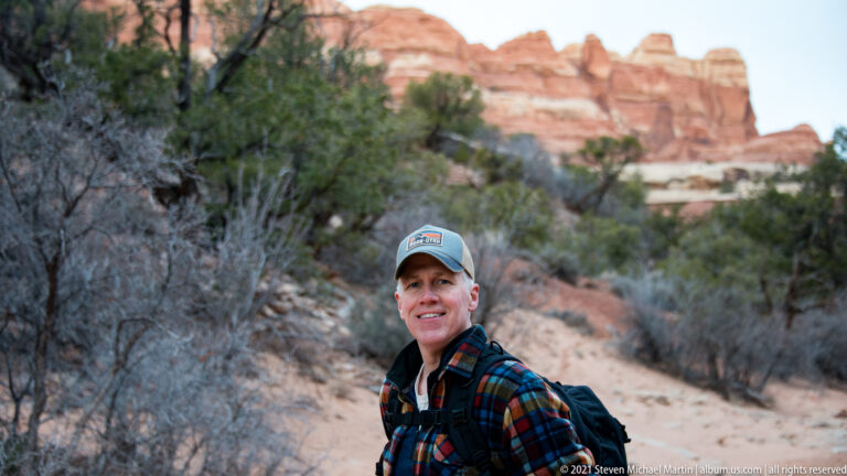A hiker in a blue cap, plaid shirt, and backpack poses before softly blurred sandstone and juniper.