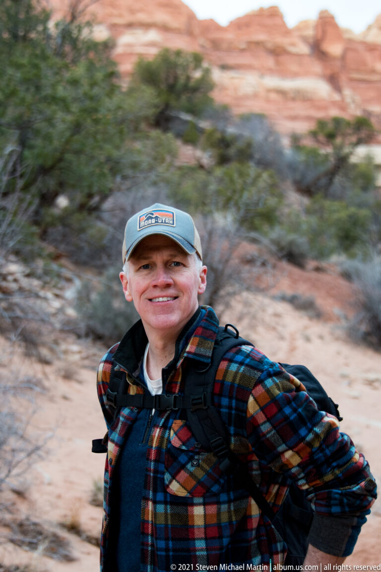 A smiling hiker in a blue cap and plaid shirt fills the frame against a blurred canyon background.