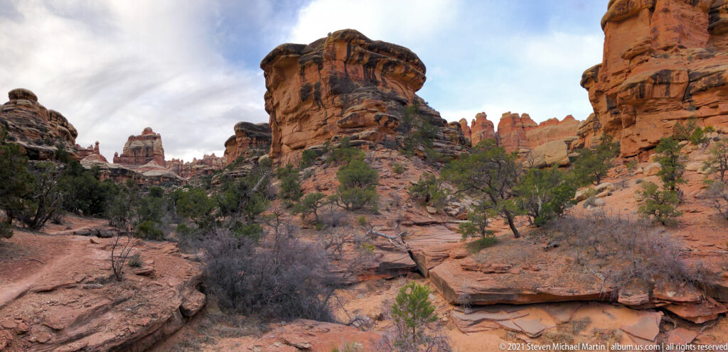 A massive banded sandstone tower stands between canyon walls above broken ledges and shrubs.