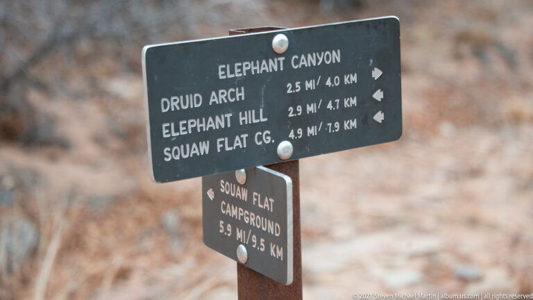 A trail sign lists Elephant Canyon, Druid Arch, Elephant Hill, and Squaw Flat Campground with distances.