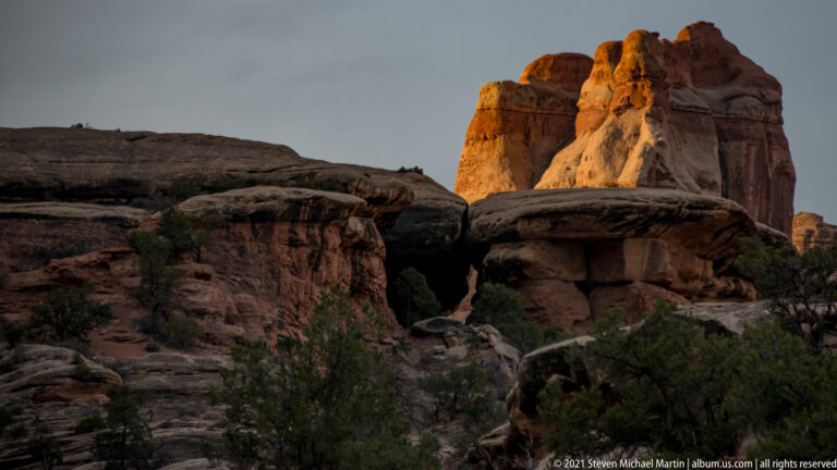 Sunlit sandstone towers rise above a shadowed foreground canyon, with sparse junipers among the rock shelves.
