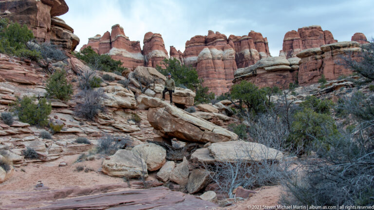 A lone visitor stands on a pale boulder below rounded red sandstone towers and scattered juniper.
