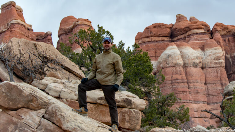A seated visitor rests on a pale boulder, with juniper and banded red sandstone rising behind him.