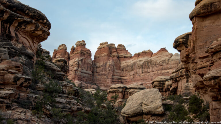 Dark sandstone walls frame a corridor opening toward distant red and cream towers, with a boulder at right.