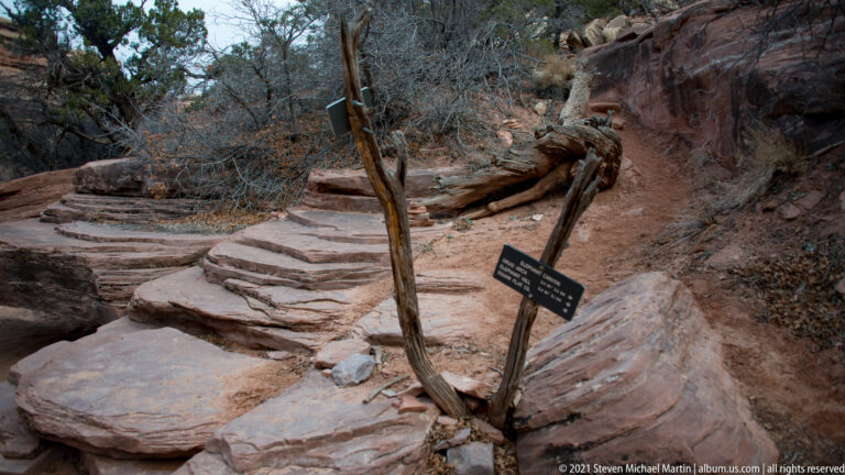 A small dark sign stands beside a sandy path among rock shelves, juniper, and leafless branches.