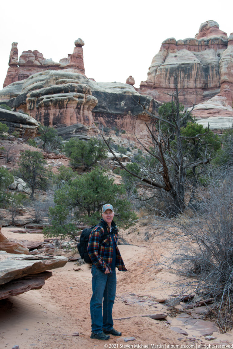A visitor stands on a sandy canyon path bordered by scrub, with banded sandstone cliffs behind.