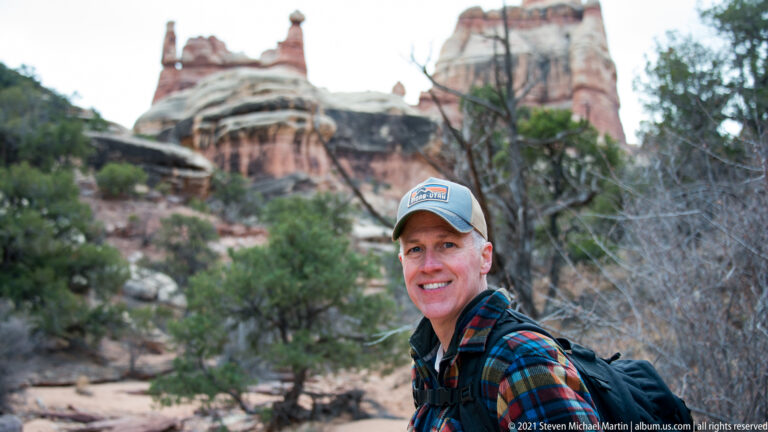 A smiling hiker in a cap and backpack stands in front of softly blurred sandstone and juniper.
