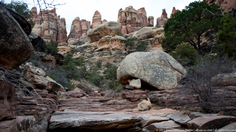 A rounded boulder rests on a smaller rock above a stepped trail, with juniper and spires behind.