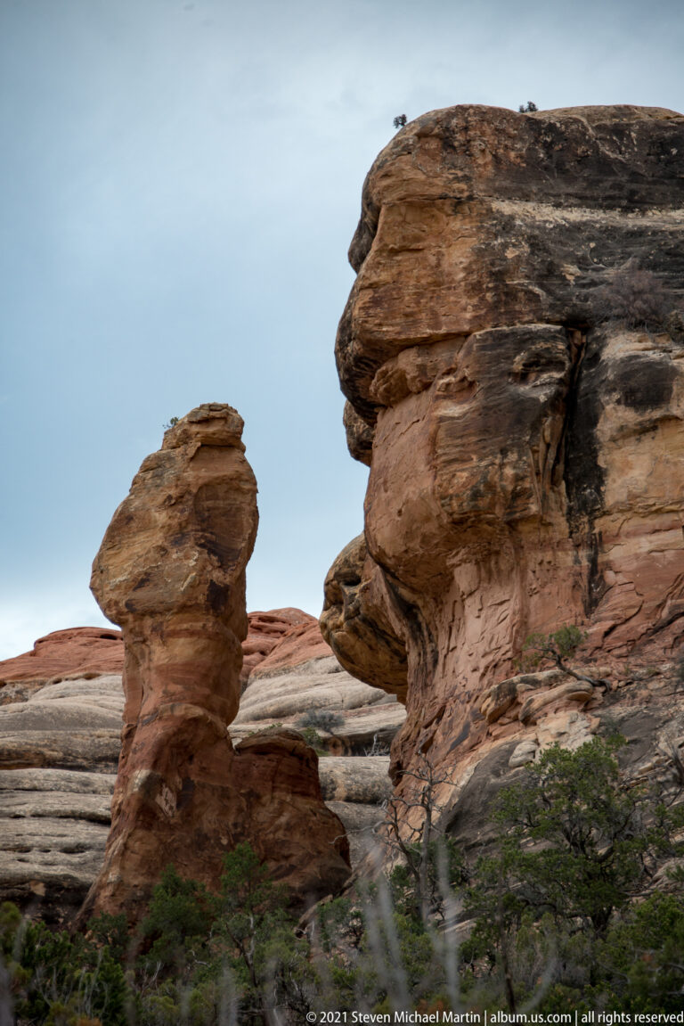 Two rounded sandstone pillars rise above horizontal ledges and low vegetation under a pale sky.