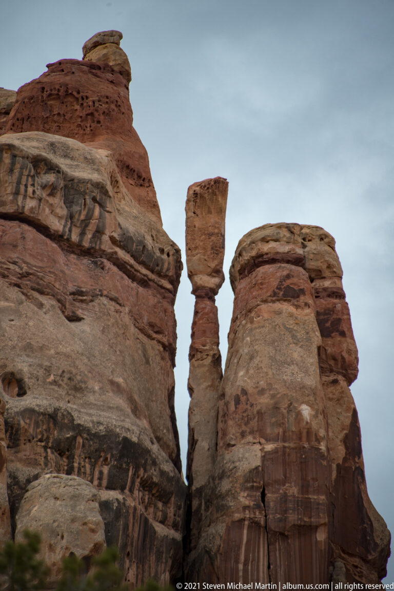 A narrow sandstone spire stands between two wider banded towers beneath a pale sky.