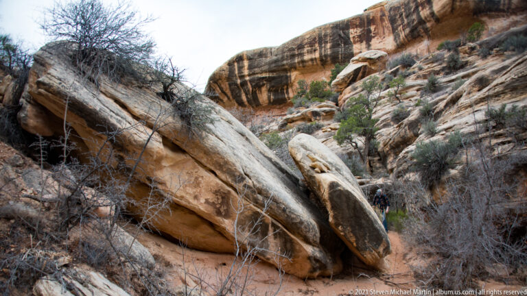 A huge tilted sandstone slab crosses the wash, with a partly obscured visitor beyond it at right.