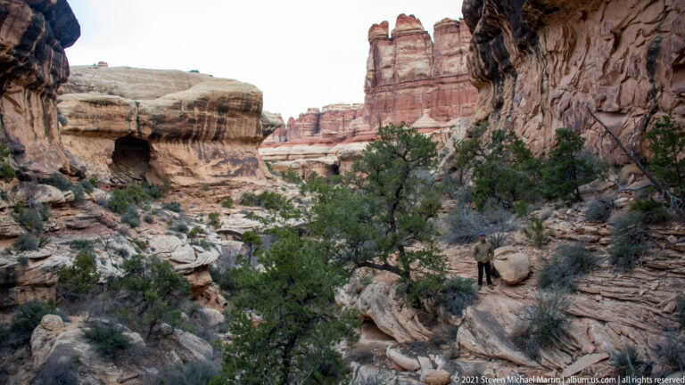 A small hiker stands among rocky ledges, with a dark alcove at left and red towers beyond.