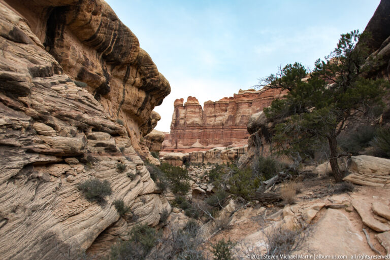 A rocky route runs between a striated sandstone wall and a juniper slope toward distant red towers.