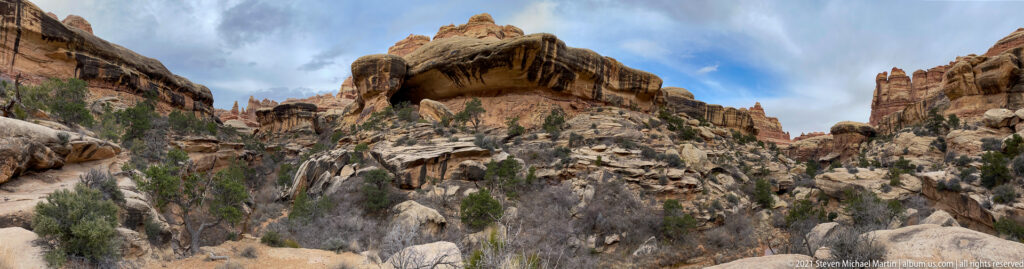 A shallow opening beneath a sandstone overhang rises above pale boulders in a narrow wash.