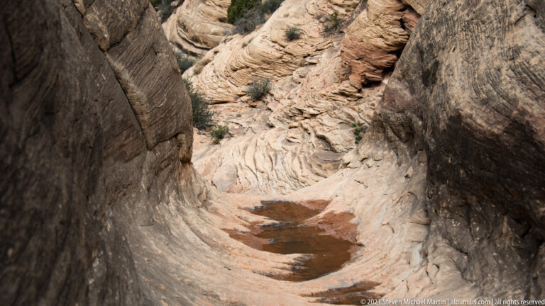 A narrow sandstone chute descends between smooth walls, with a small pool in the channel.