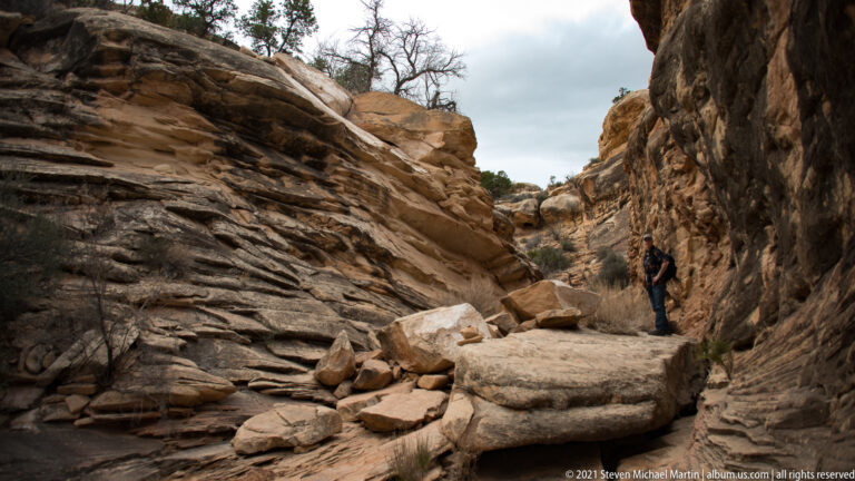 A visitor stands on a narrow ledge beside a high sandstone wall above broken rock steps.