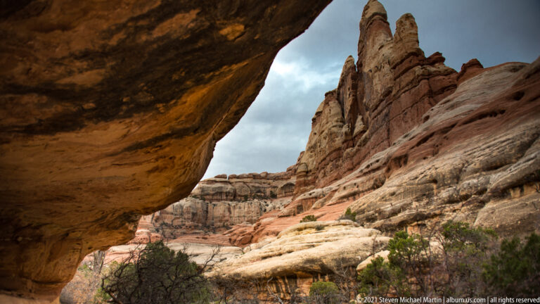 A dark overhang frames sandstone pinnacles at right and a bright corridor of layered rock beyond.