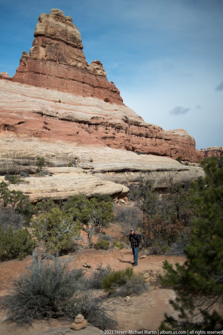 A small hiker crosses a scrub-covered basin below wide bands of pale and red sandstone.