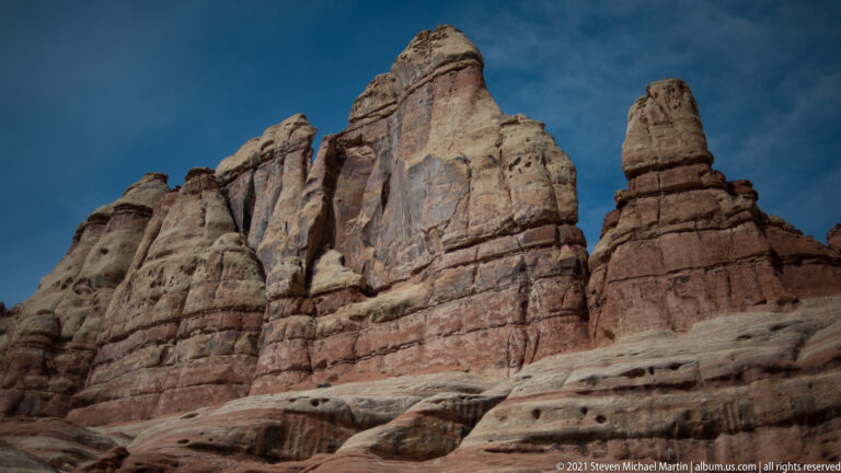 Tall red, cream, and gray sandstone spires rise against blue sky, with a separate narrow pillar at right.