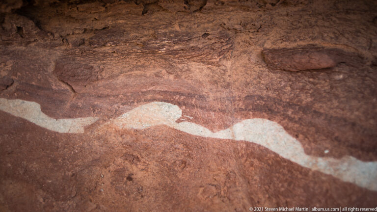 A pale wavy band runs across a close view of red sandstone with darker patches and granular texture.