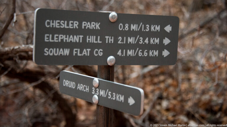 A metal trail sign lists distances to Chesler Park, Elephant Hill, Squaw Flat Campground, and Druid Arch.