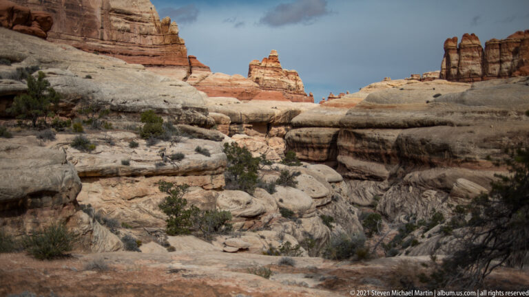 Pale sandstone shelves and a shallow drainage lead toward red rock towers beneath a partly cloudy sky.