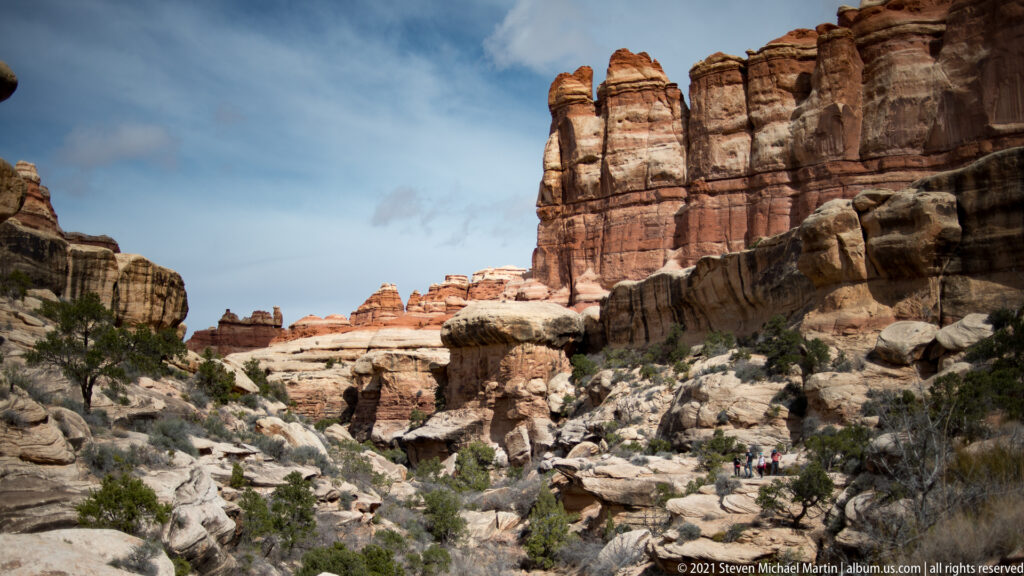 A rocky drainage winds toward red sandstone towers beneath a high layered wall and patches of blue sky.