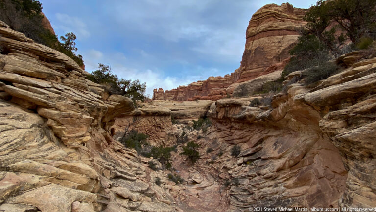 A rocky wash passes between sloping sandstone shelves and shrubs toward a banded tower.